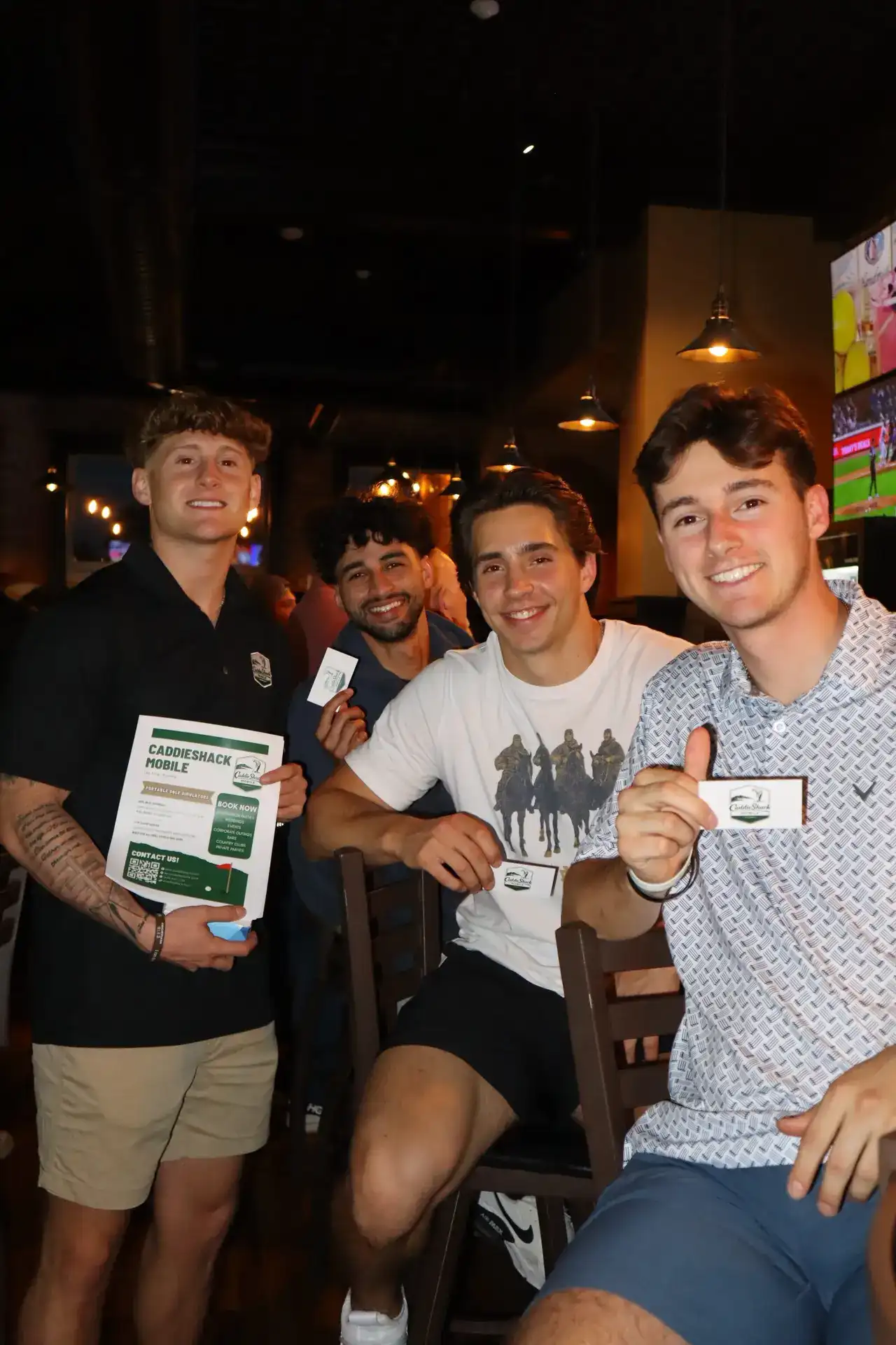 Four young men smiling with CaddieShack Mobile golf simulator flyer and business cards at a bar