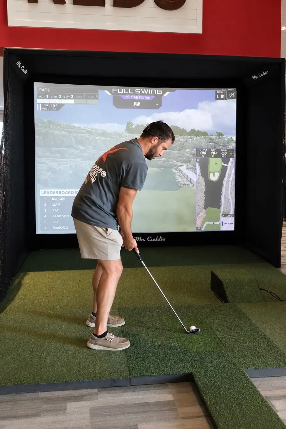 Man hitting wedge shot into a Mr. Caddie portable golf simulator screen indoors