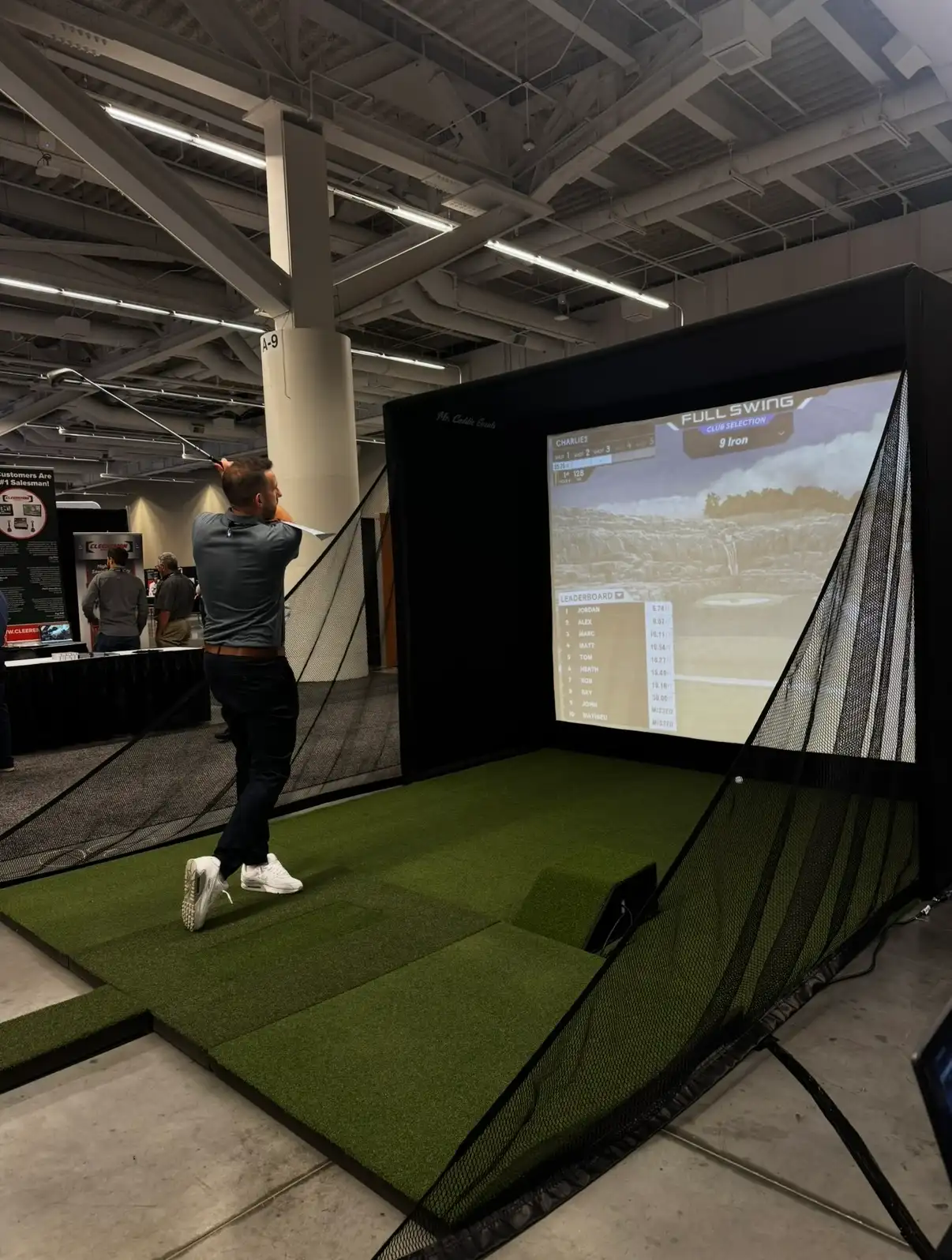 Guest mid-swing on a Mr. Caddie Events golf simulator with a live leaderboard on screen at a trade show